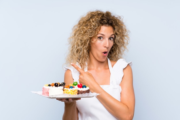 Young blonde woman with curly hair holding lots of different mini cakes surprised and pointing side