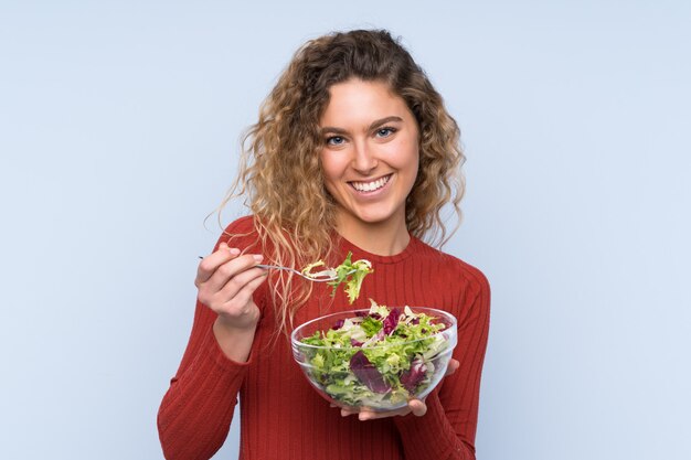 Giovane donna bionda con i capelli ricci in possesso di un insalata sul muro