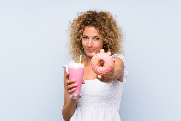 Young blonde woman with curly hair holding a strawberry milkshake and a donut