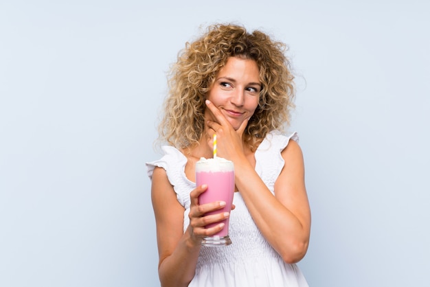 Young blonde woman with curly hair holding a strawberry milkshake thinking an idea