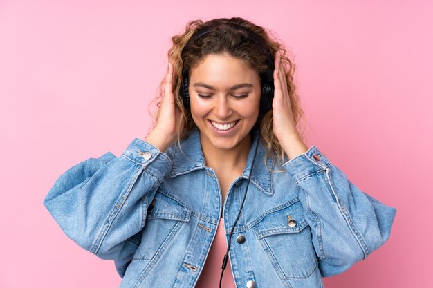 Young blonde woman with curly hair isolated on pink wall listening music