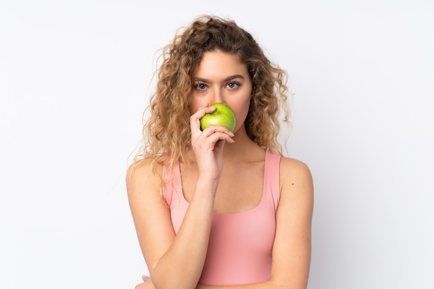 Young blonde woman with curly hair isolated on white wall eating an apple