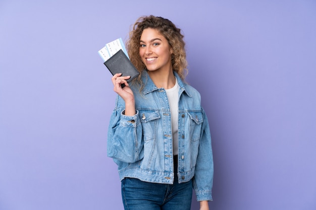 Молодая белокурая женщина с вьющимися волосами на фиолетовой стене счастлива в отпуске с паспортом и билетами на самолет