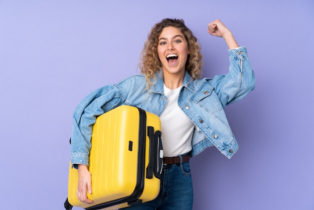 Young blonde woman with curly hair on purple wall in vacation with travel suitcase