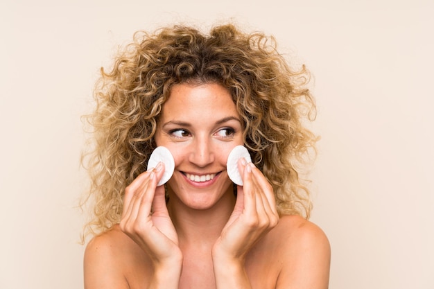 Young blonde woman with curly hair removing makeup from her face with cotton pad