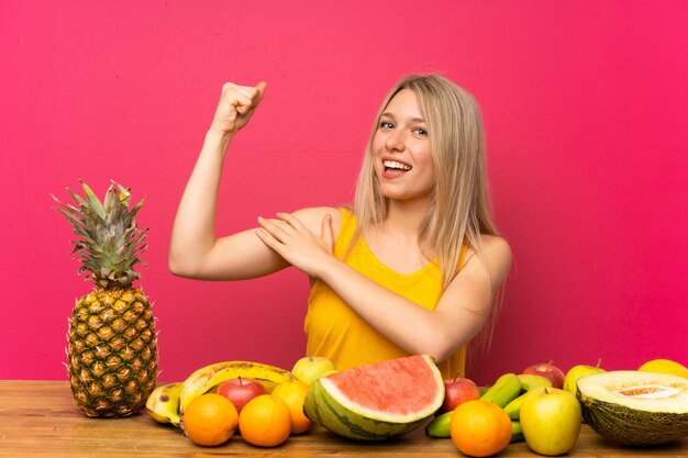 Young blonde woman with lots of fruits making strong gesture