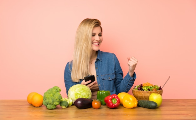 勝利の位置に携帯電話で多くの野菜を持つ若いブロンドの女性