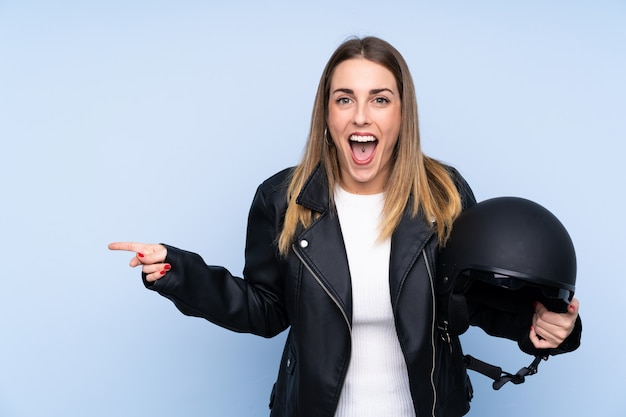 고립 된 파란색 벽 위에 오토바이 헬멧을 가진 젊은 금발의 여자가 놀라서 측면을 가리키는 손가락