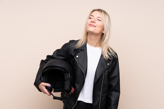 Young blonde woman with a motorcycle helmet over isolated wall looking up while smiling