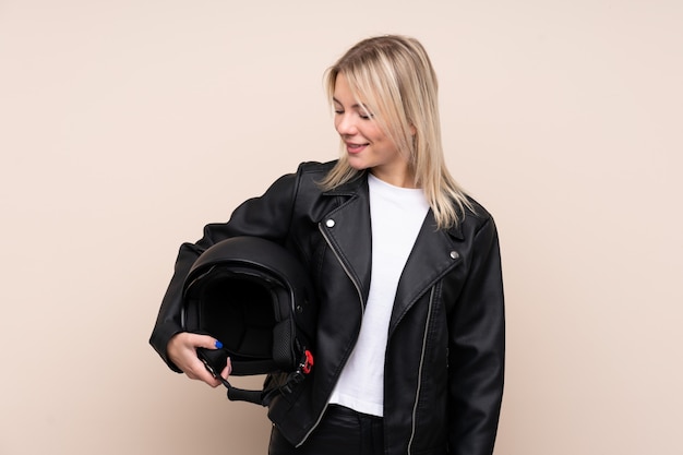 Young blonde woman with a motorcycle helmet over isolated wall with happy expression