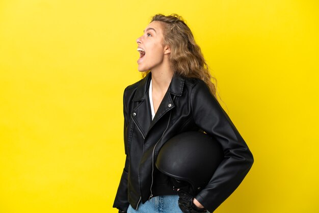 Photo young blonde woman with a motorcycle helmet isolated on yellow background laughing in lateral position