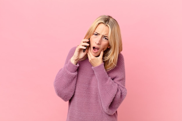 Young blonde woman with mouth and eyes wide open and hand on chin and talking with a smartphone