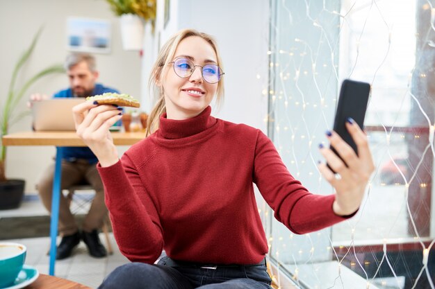 Young blonde woman with sandwich making selfie while sitting in cafe at leisure