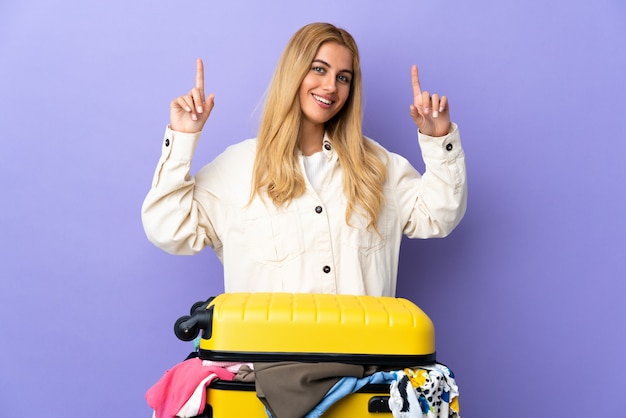 Young blonde woman with a suitcase full of clothes over isolated purple wall pointing up a great idea