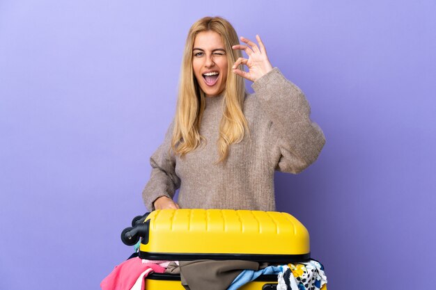 손가락으로 확인 표시를 보여주는 격리 된 보라색 벽 위에 옷의 전체 가방을 가진 젊은 금발의 여자