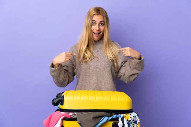 Young blonde woman with a suitcase full of clothes on isolated purple wall with surprise facial expression