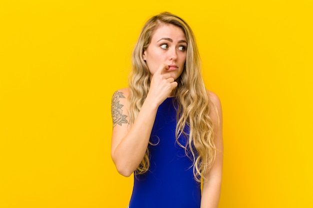 Young blonde woman with surprised, nervous, worried or frightened look, looking to the side towards copy space against yellow wall