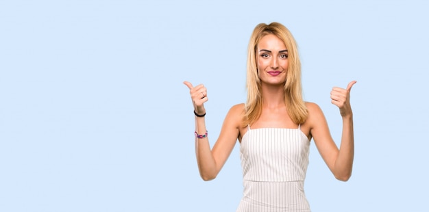 Young blonde woman with thumbs up gesture and smiling over isolated blue 