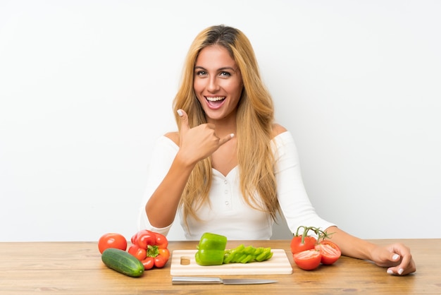 Молодая белокурая женщина с овощами в таблице делая жест телефона