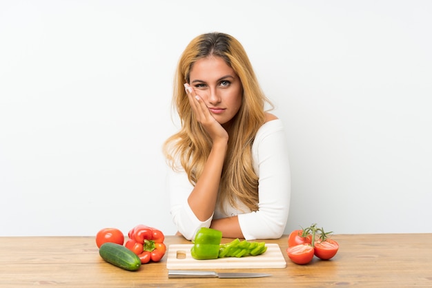 Young blonde woman with vegetables in a table unhappy and frustrated