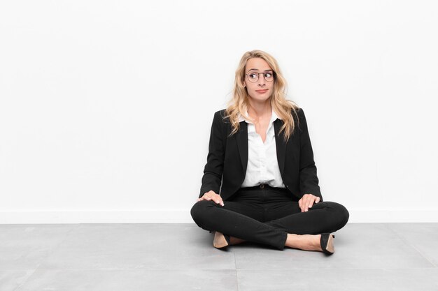 Young blonde woman wondering, thinking happy thoughts and ideas, daydreaming, looking to copy space on side sitting on the floor