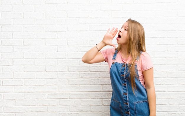 Young blonde woman yelling loudly and angrily to copy space on the side, with hand next to mouth on brick wall