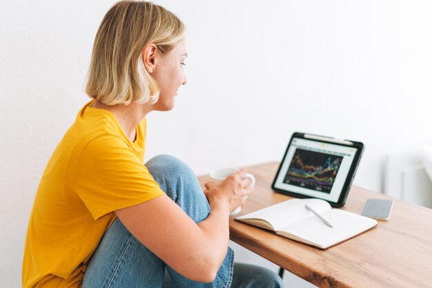 Молодая блондинка в желтой футболке изучает графики криптовалют и котировки акций на цифровом планшете