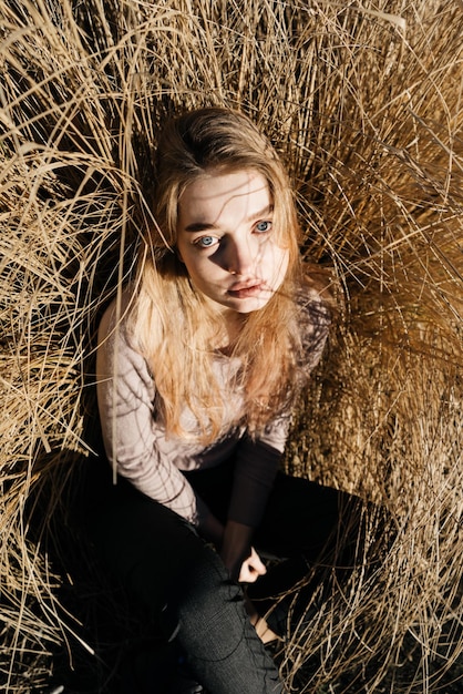 Young blue-eyed girl sits in a field in the grass, in the sun and looks at the camera