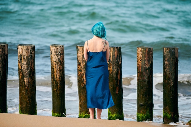 Young blue-haired woman in long dark blue dress with cute tattoo on her shoulder blade standing on sandy beach looking at sea horizon, warm coastal waves caress her feet. Freedom concept