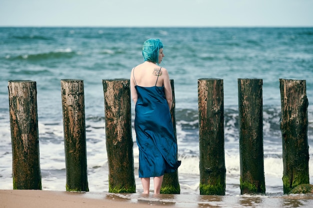 Young blue-haired woman in long dark blue dress with cute tattoo on her shoulder blade standing on sandy beach looking at sea horizon, warm coastal waves caress her feet. Freedom concept