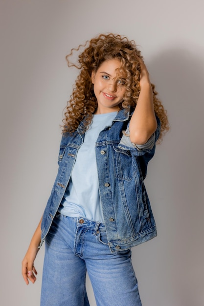 Young blueeyed blonde woman with curly hair in a denim jacket and jeans stands on a gray background