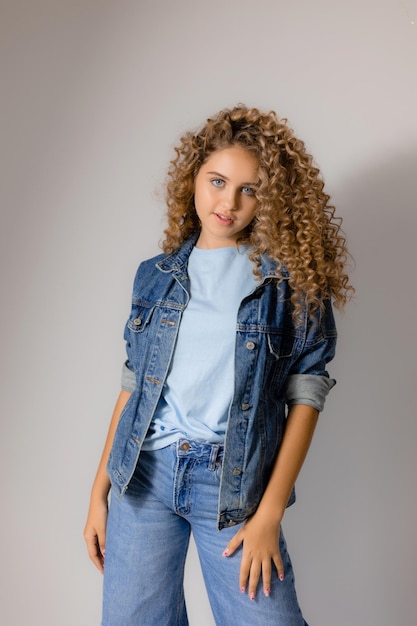 Young blueeyed blonde woman with curly hair in a denim jacket and jeans stands on a gray background