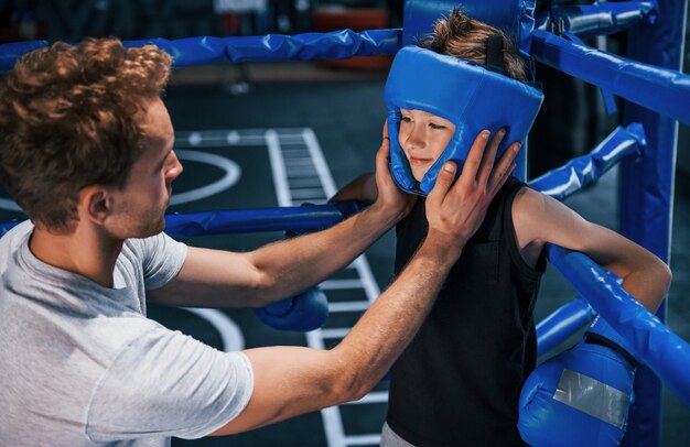 Young boxing coach is helping little boy in protective wear on the ring between the rounds.