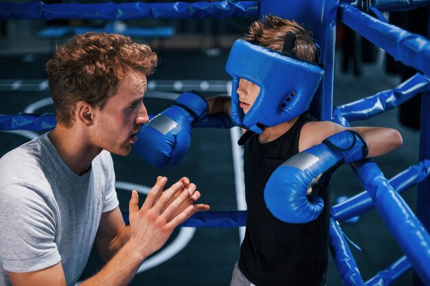 Young boxing coach is helping little boy in protective wear on the ring between the rounds.