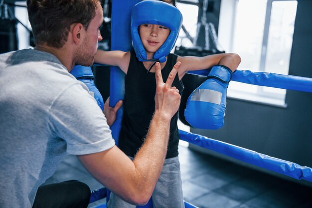 Young boxing coach is helping little boy in protective wear on the ring between the rounds.