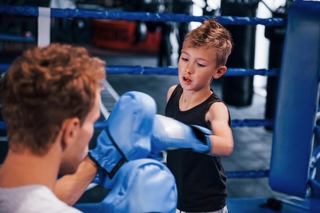 Young boxing coach is helping little boy in protective wear on
the ring between the rounds.