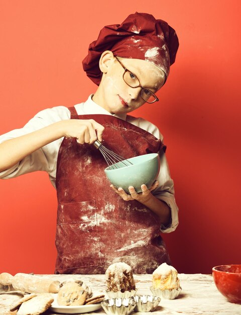 テーブルの近くに立って、赤いスタジオの背景にターコイズブルーのボウルでキッチン泡立て器で調理するガラスとステンドグラスの顔の小麦粉に制服と帽子の若い男の子かわいい料理人シェフ