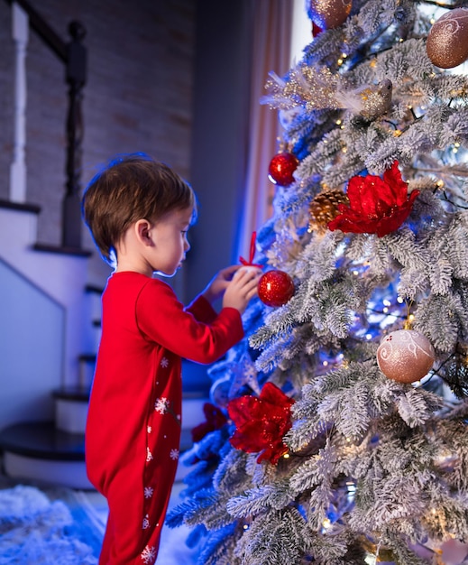クリスマスツリーに赤い装飾を飾る若い男の子 祭りのパジャマを着た若い男児が家で美しく装飾されたクリスマスツリースに赤い飾りを慎重に掛けます