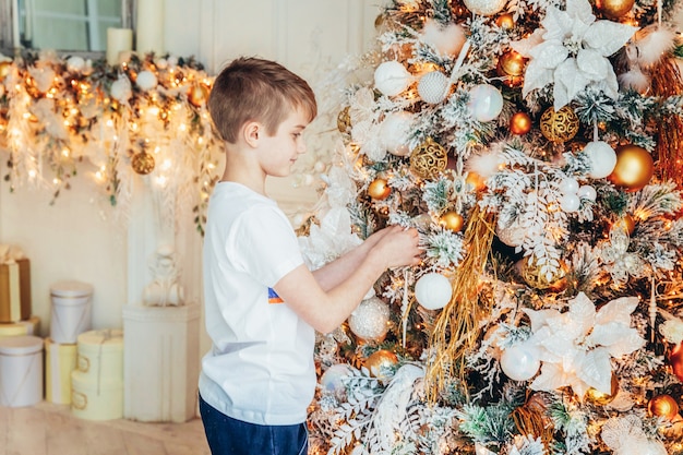 Young boy decorating Christmas tree on Christmas eve at home. Young kid in light bedroom with winter decoration. Happy family at home. Christmas New Year december time for celebration concept.