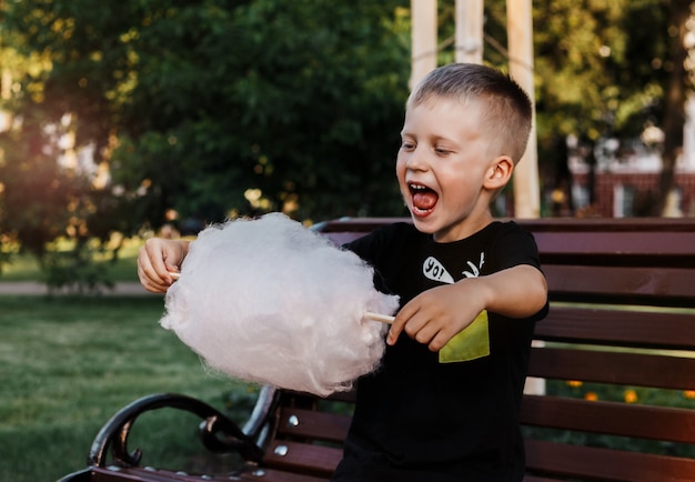 Young boy eats with pleasure cotton candy made from sticky spun sugar sitting on a bench in the park