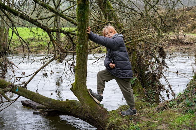 A young boy on a fallen tree near the river A boy with long hair in a warm jacket is nine years old