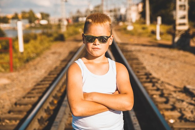 A young boy in jeans and sunglasses stands on the railroad