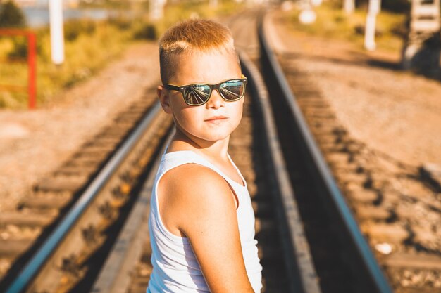 A young boy in jeans and sunglasses stands on the railroad