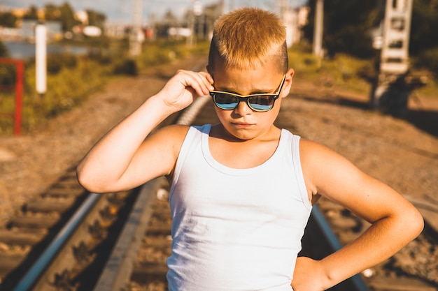 A young boy in jeans and sunglasses stands on the railroad