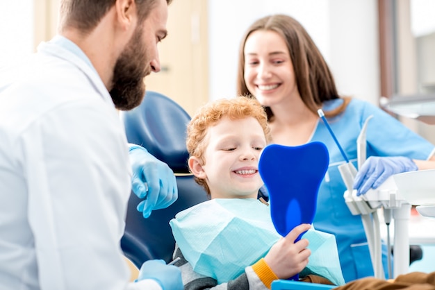 Young boy looking at the mirror with toothy smile sitting on the chair with dentist and assistant at the dental office