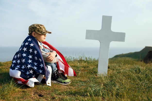 A young boy in a military cap, covered by the flag of the united states sitting on the grave of his deceased father. May 27th memorial day