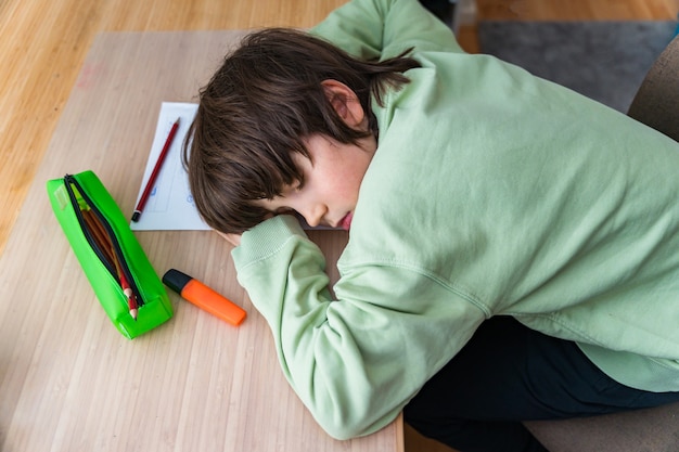 Мальчик десяти лет делает домашнее задание, сидя за столом у себя дома. усталый ребенок спит, когда делает школьные упражнения лицом на парту.