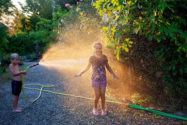 Foto un ragazzino giocosamente spruzza sua sorella con un tubo avvolgendola in una nebbia di gocce d'acqua come