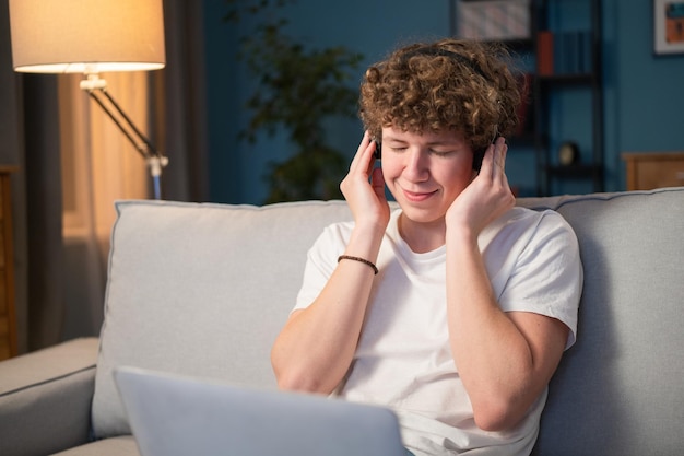 A young boy sits on a couch in the living room with a laptop on his lap and wireless headphones