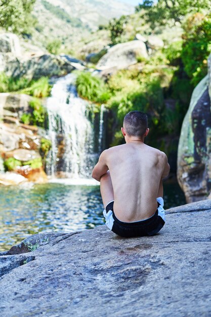 Photo young boy sitting on his back on a rock looking at the river with waterfall.